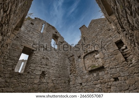 Wide angle low angle view looking up at the stone walls inside the roofless ruins of a medieval building. Historic limestone masonry of Penmon Priory with open window frames against a blue sky with wispy clouds on the Isle of Anglesey, North Wales.