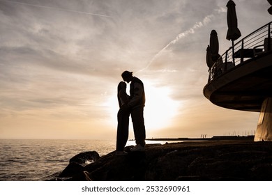 wide angle panoramic photo in backlight with silhouette of a young couple of engaged couples embracing while standing on the rocks of the sea at sunset - Powered by Shutterstock