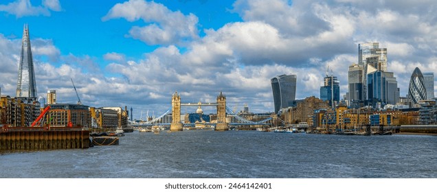Wide angle panoramic view from the River Thames of the London skyline with skyscrapers and Tower Bridge illuminated by the morning sun and boats and ferries moored at their docks.