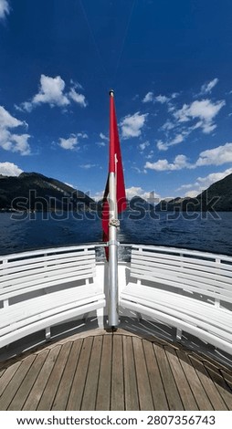 A wide angle perspective view from the aft deck of a passenger boat, looking out over a calm Swiss lake towards towering mountains.