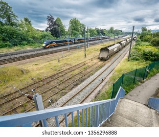 Wide Angle Photo Of High Speed Train Approaching Dickie Bridge In York Uk With Cargo Train Parked In The Sidings. Also In Photo Are Overhead Power Lines, Trees, Bridge Railings, Steps And An Alleyway