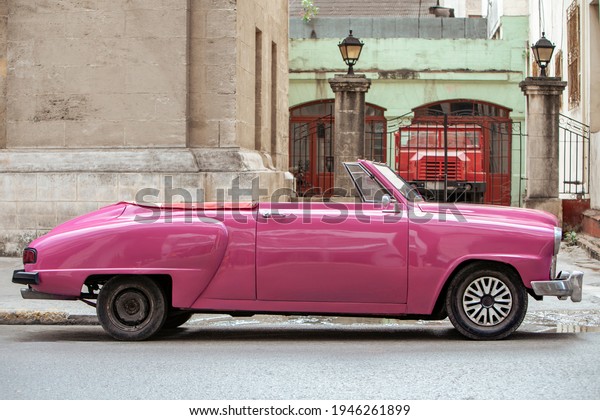 Wide angle photo of a pink convertible old American\
car parked in the streets of Havana, Cuba. Red big truck, iron bars\
and building in  background. Pavement in foreground. Saturday,\
March 24, 2018