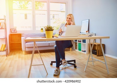 Wide Angle Picture Of Happy Young Woman Working From Her Home Office Desk With Laptop, Drinking Coffee