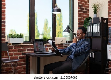 Wide Angle Portrait Of African American Startup Owner With Glasses Analyzing Marketing Indicators On Clipboard. Entrepreneur Comparing Bussiness Data With Sales Results At Desk With Laptop.