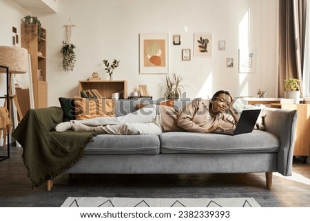 Wide angle portrait of young Black woman lying on comfortable couch in cozy home interior and using laptop, copy space
