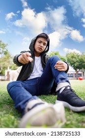 Wide angle shot of asian teenager sitting in lawn green grass pointing finger and look at camera in the park blue sky background, lifestyle portrait concept,