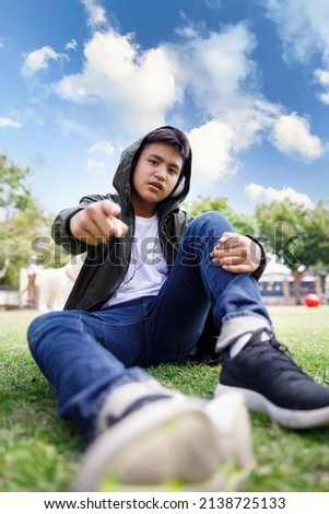 Wide angle shot of asian teenager sitting in lawn green grass pointing finger and look at camera in the park blue sky background, lifestyle portrait concept,
