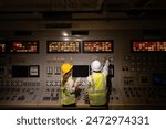 Wide angle shot in the control room of a power plant with male and female engineers working intently and pointing at buttons on the control panel of machines used to produce electricity for export.