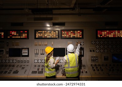 Wide angle shot in the control room of a power plant with male and female engineers working intently and pointing at buttons on the control panel of machines used to produce electricity for export. - Powered by Shutterstock