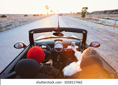 Wide angle shot of couple of teenagers or fashion internet influencers drive in cabriolet convertible mini car into sunset on palm alley. Teenage dream, forever young and happiness concept in summer