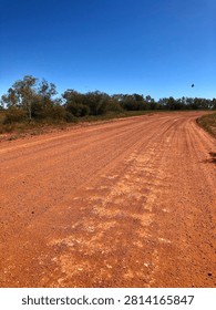 Wide angle shot of an empty red dirt desert road stretching toward the horizon under a clear blue sky with no clouds. A lonely bird of prey flies in the far distance over the rugged and remote outback landscape.