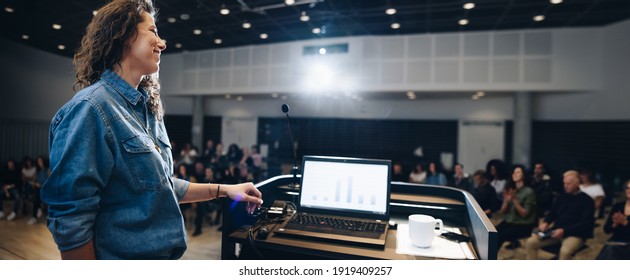 Wide Angle Shot Of A Female Speaker Standing At Podium With Laptop On The Lectern And People Sitting In The Auditorium. Businesswoman Giving A Presentation In A Corporate Event.