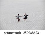 Wide angle shot of fit senior man and woman doing partner exercise balancing in seated position holding hands in middle of large tiled gray square at city park, copy space