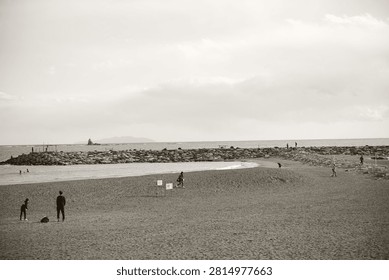 A wide angle shot of a sandy beach in Japan with stone breakwaters and several people walking and relaxing under a cloudy sky in a monochromatic sepia tone.
