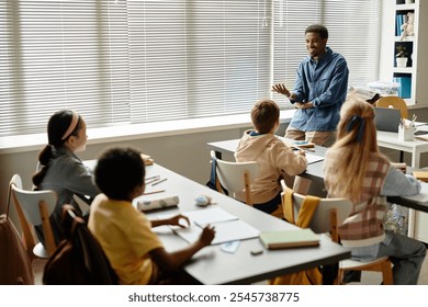 Wide angle shot of smiling male African American teacher talking to children in classroom with window blinds while explaining new topic at primary school, copy space - Powered by Shutterstock