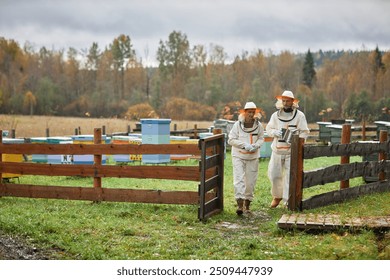 Wide angle shot of two mature beekeepers in protective suits passing through wooden farm gate leaving apiary with autumn forest on background, copy space - Powered by Shutterstock