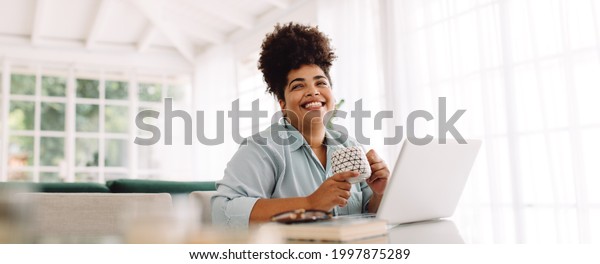 Wide angle\
shot of woman sitting on table with coffee mug and laptop. Woman\
taking break while working from\
home.