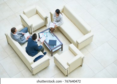 Wide Angle Top View At  Group Of Successful Business People Working Together In Hall Of Modern Office Building Sitting On Leather Sofas Around Coffee Table During Meeting.