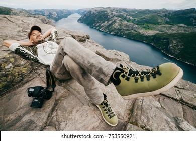 Wide Angle Unique Portrait Of Traveler. Young Man Relaxing On Stones In Norway. Guy Sleeping Outdoor. Pulplit Rock Landscape. Unusual Odd Person Lying At Edge Of Mountain With Fjords On Background