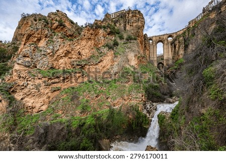 Wide angle view from the bottom of the gorge featuring the massive arches of Puente Nuevo bridge and a small waterfall in Ronda, Spain.