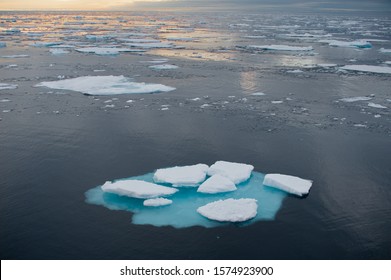 Wide Angle View With Broken Iceberg In Foreground Drifting Amongst Melting Ice Floes In Northern Arctic Ocean Viewed From Sea.A Setting Sun Casts An Orange Glow On The Sea.Climate Crisis And Emergency