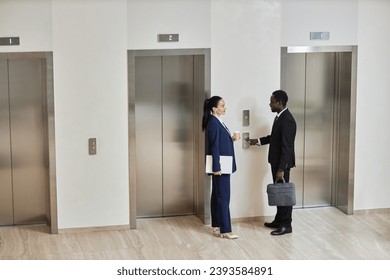 Wide angle view at Caucasian businesswoman with cup and laptop talking to cheerful African American businessman with phone and briefcase dressed in suits standing by elevators in lobby, copy space - Powered by Shutterstock