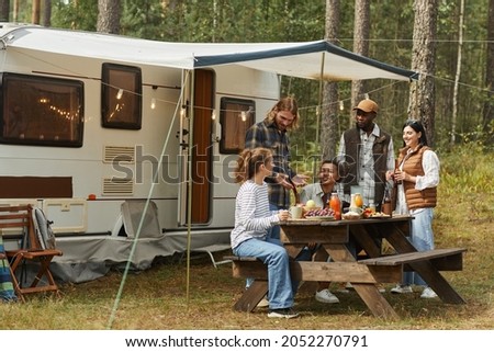 Wide angle view at diverse group of young people enjoying picnic outdoors while camping with trailer van, copy space