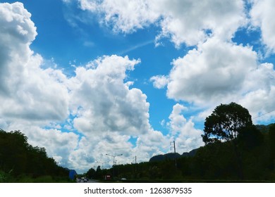 Wide angle view of Dramatic fluffy ornamental clouds covering blue sky above mountains, rain forest & rural street. Fantastic white Cumulus cloud on clear summer sky background over road, copy space