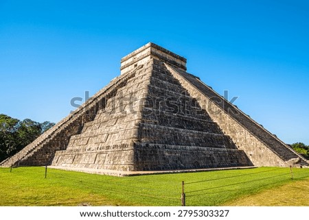 Wide angle view of the El Castillo pyramid, also known as the Temple of Kukulkan, at the Mayan archaeological site of Chichen Itza in Yucatan, Mexico. The stepped stone pyramid rises from a green grassy foreground against a clear blue sky with no people visible, emphasizing the ancient architecture and historic landmark.