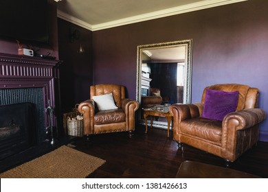 Wide Angle View Of The Interior Of A Living Room. The Walls Are Purple And There Are Two Brown Leather Arm Chairs Side By Side.