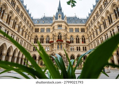 Wide angle view from a low perspective of the inner courtyard of Vienna City Hall, also known as Rathaus Vienna, in Vienna, Austria. The neo-gothic facade with arches, arcades, windows, statues, balconies and central tower is framed by green plants and an urban garden in the foreground, showing a historic government building and popular tourist attraction in the city center.