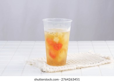 Wide angle view of a mixed fruit cocktail in a clear plastic cup set on a white rustic coaster on a table, studio setting.