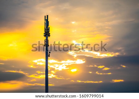 A wide angle view of a monopole cell site tower at sunset. Golden backlit silhouetted cellular base station with scattered clouds and copy space