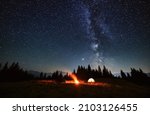 Wide angle view on beautiful landscape in the mountains. Tourist tents and campfire under beautiful starry sky. Mountains range behind spruces. Concept of night camping and astrophotography