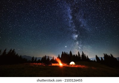 Wide angle view on beautiful landscape in the mountains. Tourist tents and campfire under beautiful starry sky. Mountains range behind spruces. Concept of night camping and astrophotography