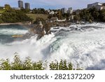 Wide angle view of a powerful cascading waterfall with rushing white foam near a distant town, Neuhausen