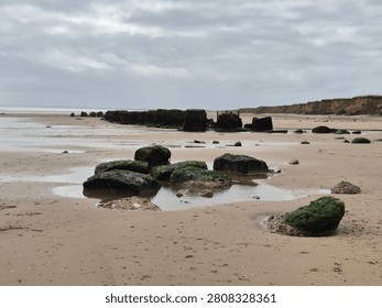 Wide Angle View Of Sandy Beach With Rock Groynes Moss Covered Stones Tidal Pools And Coastal Cliff Line Under Overcast Sky Conditions In Daylight 