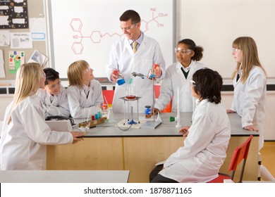 A Wide Angle View Of A Science Laboratory With Students In Lab Coats And Safety Goggles As They Are Watching An Experiment Conducted By The Chemistry Teacher