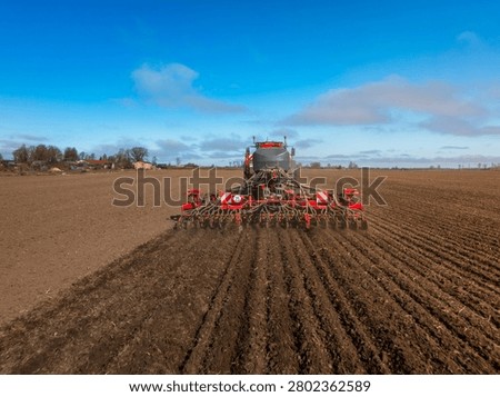 Wide angle view shows a tractor towing a seed drill cutting parallel furrows. Seed tubes and coulters are visible. Rural field, cool clear day, strong daylight.