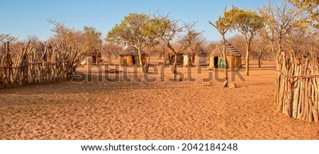 Wide angle view of a small Himba village in rural Namibia, with traditional round mud houses, thatched roofs and wooden goat pens in a sandy, arid landscape.