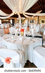 A Wide Angle View Of A Wedding Reception Venue, With Dance Floor  And Surrounding Tables.  A Large Chandelier In The Middle Of The Room Has Tooling Draped From It, With Mini Lights.