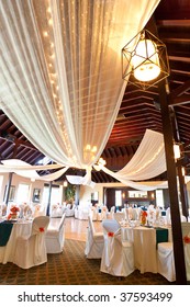 A Wide Angle View Of A Wedding Reception Venue, With Dance Floor And Surrounding Tables.  A Large Chandelier In The Middle Of The Room Has Tooling Draped From It, With Mini Lights.