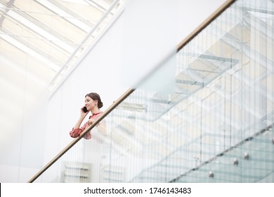 Wide Angle View At Young Businesswoman Speaking By Smartphone While Standing On Balcony In Minimal Office Building Interior, Copy Space