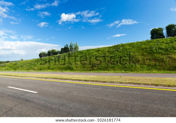 wide asphalt\
road divided into several lanes, around the road there are hills\
with green grass, summer\
landscape