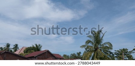 A wide atmospheric composition captures a typical Indonesian rural village scene: traditional brick houses with distinctive red corrugated metal rooftops, flanked by tall coconut palms beneath a dramatic blue sky with sweeping white cloud formations. The image conveys the quiet beauty of everyday Indonesian life — red roofs, green palms, and endless sky — projecting authenticity, warmth, and cultural identity. The generous sky with dynamic clouds provides natural copy space, while the earthly lower portion grounds the scene in real rural Indonesian life. Useful for travel editorial content, Indonesia tourism promotion, cultural documentary projects, eco-village lifestyle campaigns, and social media backgrounds showcasing local community life. The blend of traditional architecture and tropical nature provides a distinctly Indonesian visual identity that resonates with global markets seeking authentic Southeast Asian imagery.