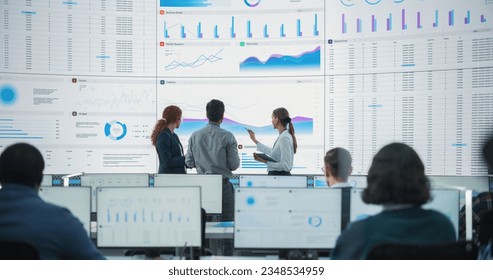 Wide Back Shot of Diverse Team of Data Scientists Discussing Graphs And Reports On Big Digital Screen In Monitoring Office. Multiethnic Employees Working On Desktop Computers For Consulting Company.