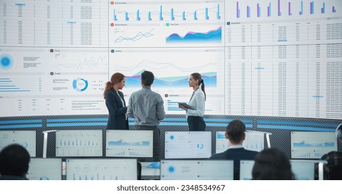 Wide Back Shot of Diverse Team of Data Scientists Discussing Graphs And Reports On Big Digital Screen In Monitoring Office. Multiethnic Employees Working On Desktop Computers For Deep Learning Company