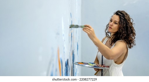 wide banner panorama portrait of sexy female beauty artist woman with curly hair in casual white underskirt with brush and palette painting on the studio wall mural, copy space