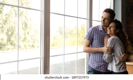Wide banner panoramic view of happy millennial Caucasian couple look in window distance dreaming or thinking. Smiling young man and woman hug embrace celebrate moving to own home or house.