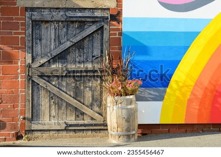 A wide black wooden door with paint fading and a barrel of flowers. The storage buildings' door has wrought iron hinges and a handle. There's a blue sky and rainbow painted on the wall of the building